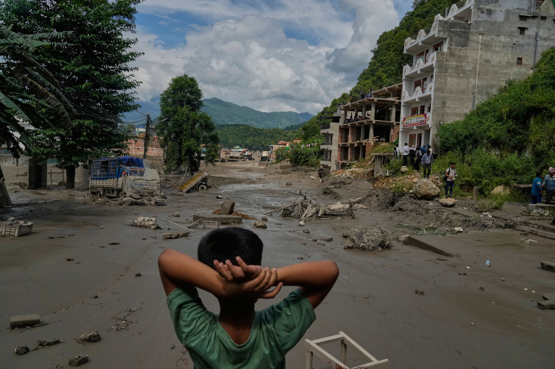 A resident looks out over a flood-devastated street in northern Nepal: thick mud and debris cover the valley, a truck is half-buried and buildings sit damaged at the water's edge